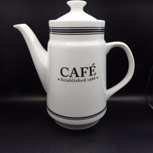 May include: White ceramic coffee pot with a black and white striped design. The pot has a black inscription that reads "CAFÉ Established 1986".