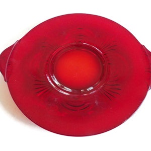 Può includere: Un piatto da portata in vetro rosso vibrante con un design circolare. Il piatto ha un'area centrale incassata e due piccole maniglie su entrambi i lati. Il design presenta linee incise che si irradiano dal centro. Un oggetto vintage per la cucina.