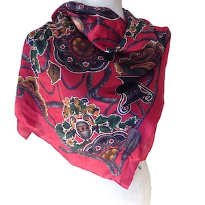 May include: A red silk scarf with a detailed pattern of nautical elements, including ropes, tassels, and decorative emblems. The scarf features accents of navy blue, gold, and green, creating a luxurious and elegant accessory.
