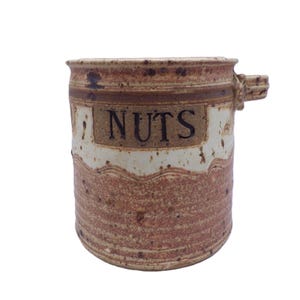 Puede incluir: Tarro de cerámica hecho a mano con un diseño rústico. El tarro es principalmente marrón con una banda color crema y la palabra "NUTS" en letras marrón oscuro. Tiene un pequeño asa en el lateral y es ideal para guardar frutos secos u otros productos secos.