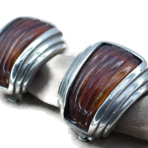 May include: A pair of silver tone clip-on earrings with amber colored glass inserts. The glass has a striated pattern.