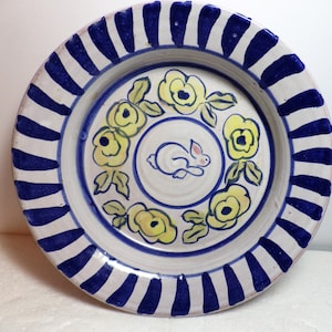 Op de afbeelding: Een wit keramiek bord met een blauw-witte gestreepte rand. Het midden van het bord heeft een wit konijn omringd door gele bloemen.