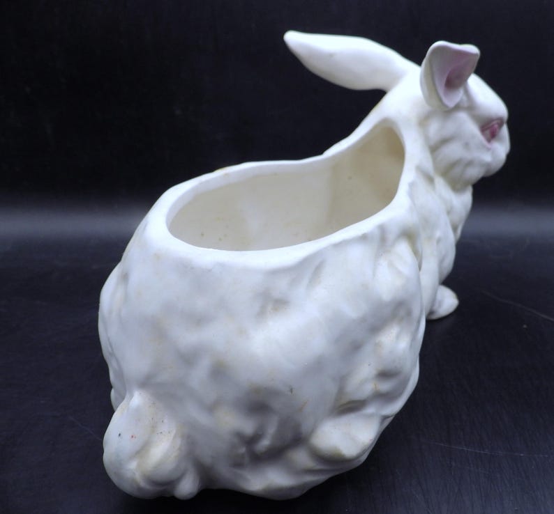 May include: A white ceramic planter shaped like a rabbit. The rabbit has pink accents on the ears and eyes. The planter has a large opening on the back for planting flowers or other plants. The planter is a decorative item.