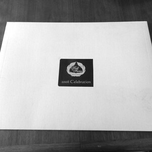 Può includere: Una carta rettangolare bianca con un quadrato nero al centro. Il quadrato presenta un logo con le parole "Hall of Fame" e il testo "2006 Celebration" sotto. La carta è su una superficie di legno.