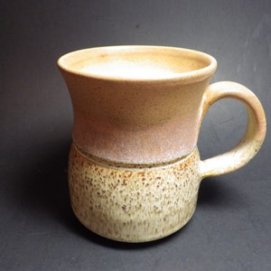 May include: A handmade ceramic mug with a brown and beige speckled glaze. The mug has a wide, rounded base and a flared lip.