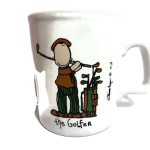 May include: White ceramic coffee mug with a cartoon illustration of a golfer with a golf bag and clubs. The text "the Golfer" is printed below the illustration. The text "William Egan © 1975" is printed on the right side of the mug.