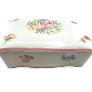 May include: A white ceramic trinket box with a pink trim and a floral design. The lid features a cluster of pink, yellow, and blue flowers with green leaves.