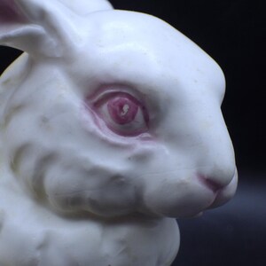 May include: A close-up of a white ceramic rabbit figurine. The rabbit has pink details around the eyes and nose. The background is a dark, solid color.