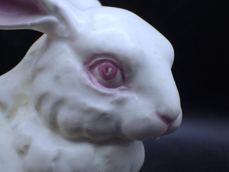 May include: A close-up of a white ceramic rabbit figurine. The rabbit has pink details around the eyes and nose. The background is a dark, solid color.