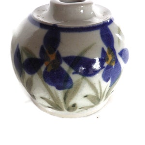 May include: A small, round ceramic vase with a light gray base and a dark blue rim. The vase is decorated with blue iris flowers with yellow centers and green leaves. The vase has a small opening at the top.
