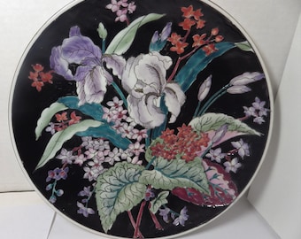 May include: A black ceramic plate with a floral design featuring purple, white, and red flowers. The flowers are painted in a realistic style and are surrounded by green leaves.