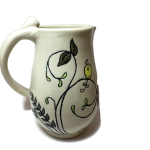 May include: A white ceramic pitcher with a whimsical design of a yellow bird perched on a green vine with leaves. The pitcher has a handle and a rounded body.