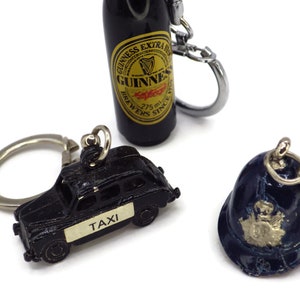 Puede incluir: Tres llaveros: un taxi londinense en miniatura negro, un casco de policía azul y una botella de cerveza Guinness en miniatura.