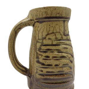 May include: A handcrafted ceramic mug with a handle. The mug features a textured, earthy design with shades of brown and green. The mug's exterior has horizontal and vertical lines, giving it a rustic appearance. The mug is suitable for hot or cold beverages.