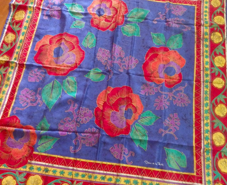 Pu&ograve; includere: Un foulard di seta blu con un motivo floreale. I fiori sono rossi, arancioni e viola con foglie verdi. Il foulard ha un bordo rosso, giallo e verde.