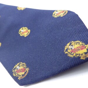 May include: A navy blue tie featuring a repeating pattern of gold and red crests. Each crest has a central emblem and text. The fabric has a textured appearance, and the crests are evenly distributed across the tie's surface.