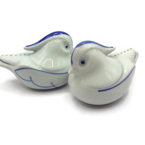 May include: Two white ceramic bird figurines with blue and gold accents. The birds are facing each other and have a rounded shape.