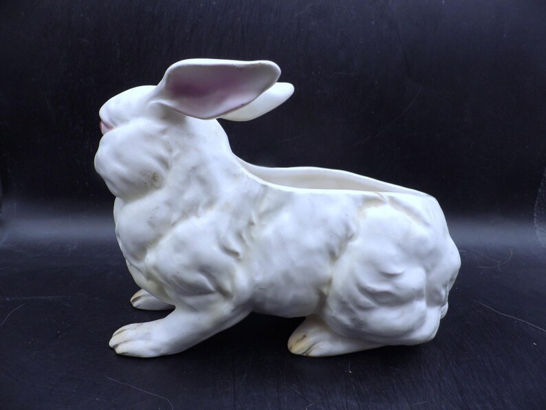 May include: A white ceramic rabbit planter with a hollowed-out back for plants. The bunny has pink accents on the ears and nose. The planter is in a sitting position, with its front paws extended.