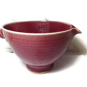 May include: A deep red ceramic mixing bowl with a pouring spout and two handles. The bowl has a textured, speckled finish and a slightly raised base.