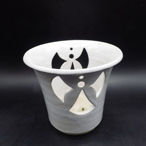 May include: A gray ceramic candle holder with a white rim and cut-out butterfly designs. The candle holder has a cylindrical shape with a slightly flared top. The cut-out designs allow light to shine through, creating a decorative effect.