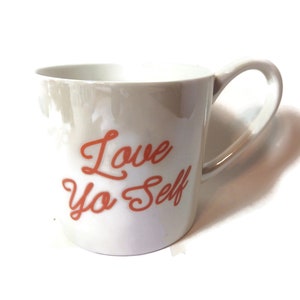 Op de afbeelding: Witte keramische mok met een rode, cursieve inscriptie die "Love Ya Self" zegt.