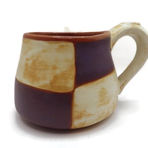 May include: Handmade ceramic mug with a unique patchwork design. The mug features alternating squares of cream, purple, and brown. The handle and rim are a lighter cream color. A perfect addition to any kitchen or coffee collection.