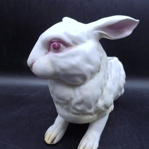 May include: A white ceramic rabbit figurine with pink inner ears and eyes. The rabbit is in a seated position, with its front paws extended. The figurine has a textured surface and is set against a dark background. The rabbit's nose and paws are a light yellow color.