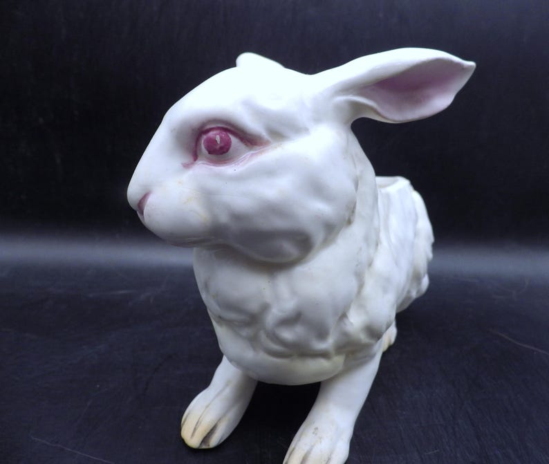 May include: A white ceramic rabbit figurine with pink inner ears and eyes. The rabbit is in a seated position, with its front paws extended. The figurine has a textured surface and is set against a dark background. The rabbit's nose and paws are a light yellow color.