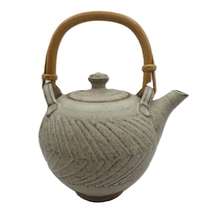 May include: A ceramic teapot with a light beige glaze and a textured, diagonal pattern. It features a bamboo handle and a small, rounded lid. The spout is short and curved.
