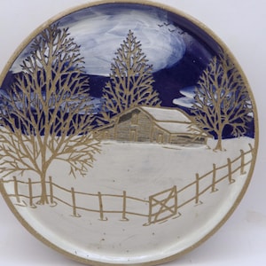 May include: A ceramic plate with a winter scene. The plate features a barn, trees, and a fence in a snowy landscape. The sky is dark blue with white clouds. The plate has a beige rim.
