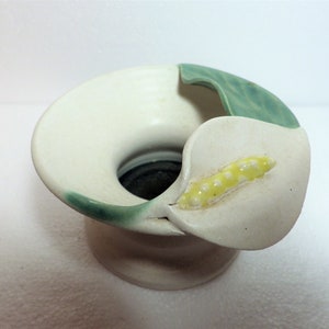 May include: White ceramic flower vase with a green leaf and a white flower with yellow accents. The vase is shaped like a flower.