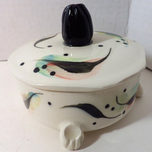 May include: A white ceramic trinket box with a black knob on the lid. The box has a whimsical design of black, green, and pink abstract shapes and black dots.