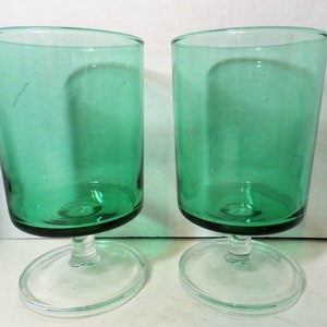 May include: Two green glass drinking glasses with clear glass stems and bases. The glasses are empty and sitting on a white surface.