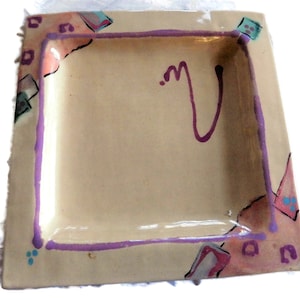 May include: A square ceramic dish with a light beige background and a purple border. The dish has a hand-painted design with pink, blue, and green accents. There is a purple squiggle in the centre of the dish.