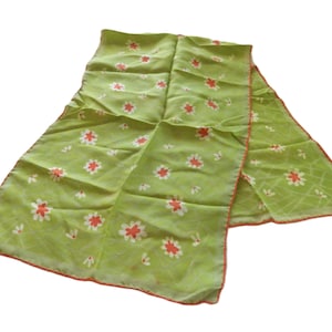 May include: A rectangular lime green scarf featuring a repeating pattern of small white and orange flowers. The fabric has a subtle grid-like texture throughout, and the edges are finished with a thin, contrasting orange border trim.