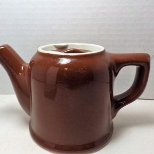 Puede incluir: Una tetera de cerámica marrón con un borde blanco y un asa blanca. La tetera es pequeña y tiene un cuerpo redondeado.
