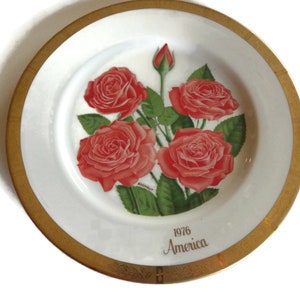 Può includere: Un piatto in porcellana bianca con un bordo dorato presenta un motivo floreale di quattro rose rosse con foglie verdi. Il piatto ha il testo "1976 America" stampato in oro sul fondo.
