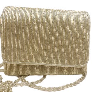May include: A rectangular, beaded handbag in a light gold color. The bag features a flap closure and a long, beaded shoulder strap. The surface is covered in vertical rows of small beads, creating a textured appearance. The bag is a fashion accessory.