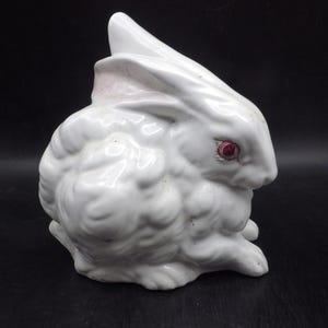 May include: A white ceramic rabbit figurine with a textured, curly fur pattern and distinct red eyes. The rabbit is depicted in a seated position with its ears pointed upward, showcasing a glossy finish and detailed sculpting across its body.