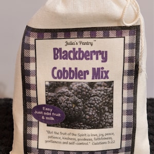 May include: A white canvas bag with a drawstring closure. The bag has a printed label that says "Julia's Pantry Blackberry Cobbler Mix". The label also includes a picture of blackberries and a quote from Galatians 5:22.