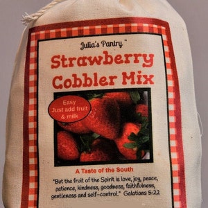 May include: A canvas bag labeled "Julia's Pantry Strawberry Cobbler Mix." The label features a red and white checkered border, a photo of strawberries, and the text "Easy Just add fruit & milk." The bag is tied with a natural rope.