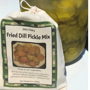 May include: A white cloth bag with a red drawstring, containing a mix for fried dill pickles. The bag has a label that reads "Julia's Pantry Fried Dill Pickle Mix" and includes a Bible verse. The bag is next to a jar of dill pickles.