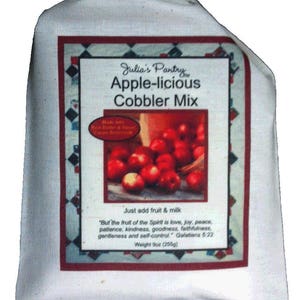 May include: A white bag with a red and white label that says "Julia's Pantry Apple-licious Cobbler Mix". The label has a picture of red apples in a bowl and a quote about the fruit of the Spirit. The bag is 9 ounces (256 grams).