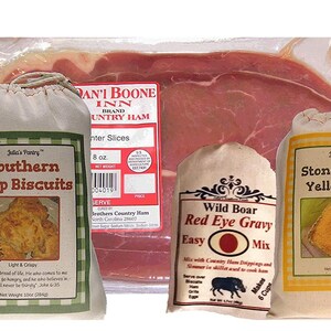 May include: Four bags of food products, including a bag of Southern Drop Biscuits, a bag of Stone Ground Yellow Grits, a bag of Wild Boar Red Eye Gravy Mix, and a package of Dan'I Boone Inn Brand Country Ham. The bags are made of white fabric with green and brown accents. The package of ham is clear plastic with a red label.