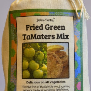 May include: A white canvas bag with a drawstring closure. The bag has a label that reads "Julia's Pantry Fried Green TaMaters Mix". The label also includes a picture of green tomatoes and a cornbread muffin. The label includes the text "Delicious on all Vegetables" and a Bible verse from Galatians 5:22. The bag is labeled with a net weight of 9 ounces (256 grams).