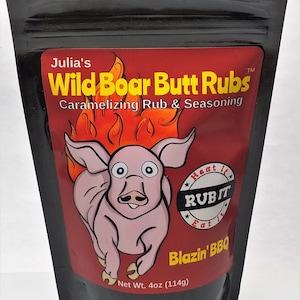 Puede incluir: Una bolsa negra resellable con una etiqueta roja que presenta un cerdo de dibujos animados corriendo con llamas. La etiqueta dice "Julia's Wild Boar Butt Rubs Caramelizing Rub & Seasoning" y "Heat It Rub It Eat It Blazin' BBQ". La bolsa contiene 114 gramos de condimento.