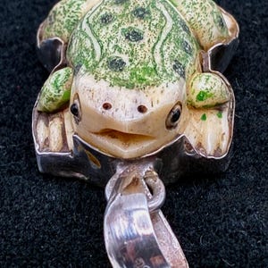 May include: A frog pendant with a detailed design. The frog has a cream-colored body with green spots and a silver-colored frame. The pendant has a loop for a chain and is marked with "925".