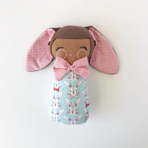 May include: A brown bunny doll with pink polka dot ears and a pink bow. The doll is wearing a blue and white fabric with a pattern of bunnies wearing bow ties.