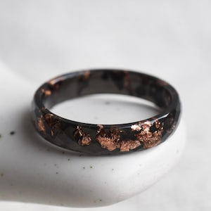 Peut inclure: Une bague noire avec des flocons de couleur cuivre incrustés. La bague est posée sur une surface blanche. La bande a un design à facettes, captant la lumière. Cette bague unique est un bijou.