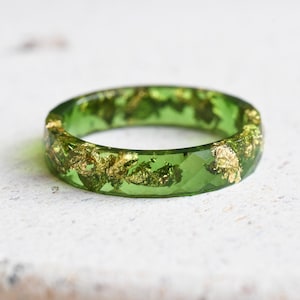 May include: A green resin ring with gold leaf inclusions. The ring has a faceted design and a clear, transparent appearance, showcasing the gold flakes suspended within the green material. The ring is a piece of jewellery.